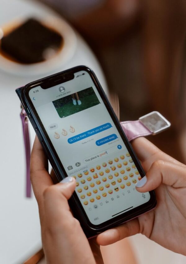 A person holds a smartphone displaying a text conversation with emojis on the screen. Their fingers are near the emoji keyboard. A plate with food is blurred in the background.