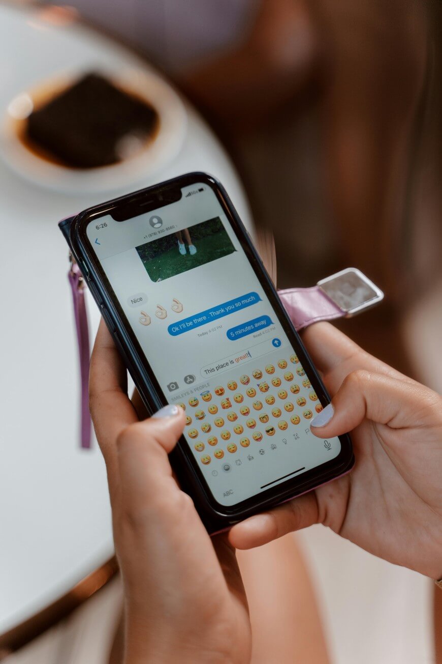 A person holding a smartphone and typing a text message with emojis. The screen displays a messaging app with a conversation and a row of emoji options. A blurred plate with food is in the background.