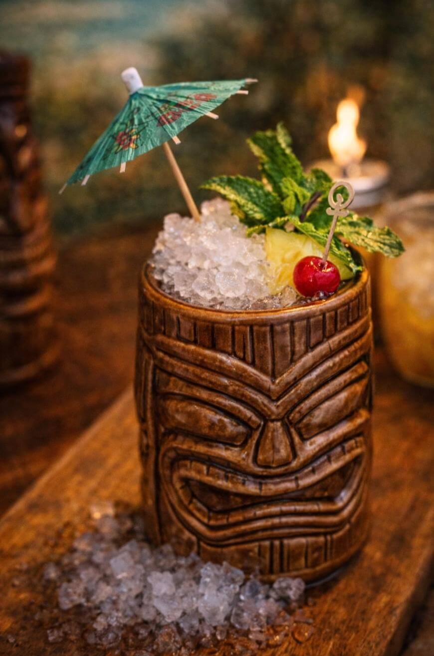 A tiki mug filled with crushed ice, garnished with a paper umbrella, mint, pineapple, and a cherry, sits on a wooden surface with some ice spilled around it.