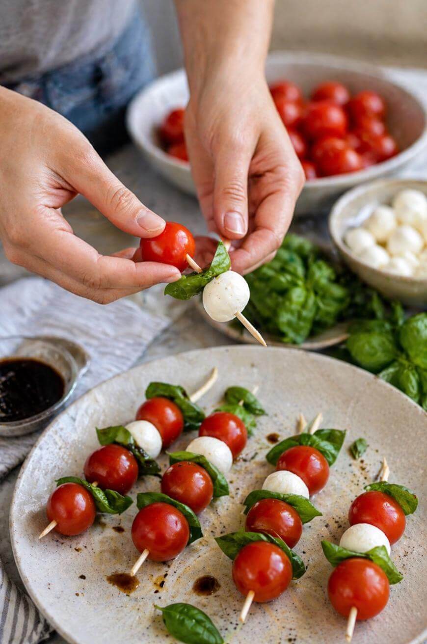 A person’s hands are assembling Caprese skewers with cherry tomatoes, mozzarella balls, and fresh basil leaves. Several completed skewers are on a plate, with bowls of ingredients and balsamic glaze nearby.