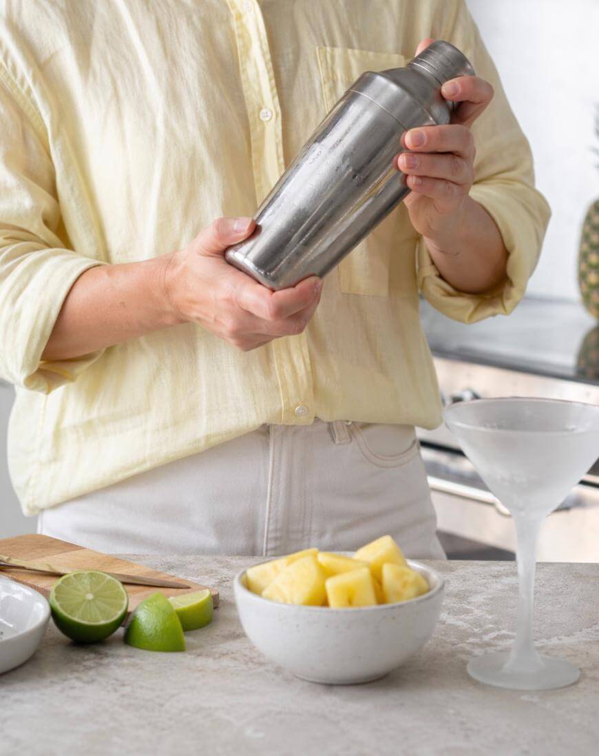 A person in a light yellow shirt holds a cocktail shaker. On the counter are a bowl of pineapple chunks, lime halves, and an empty martini glass. The setting appears to be a bright kitchen.