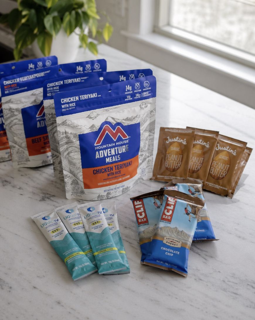 Assorted packaged camping and adventure snacks on a marble counter, including Mountain House Adventure Meals, Justin’s peanut butter packets, CLIF bars, and protein drink tubes.