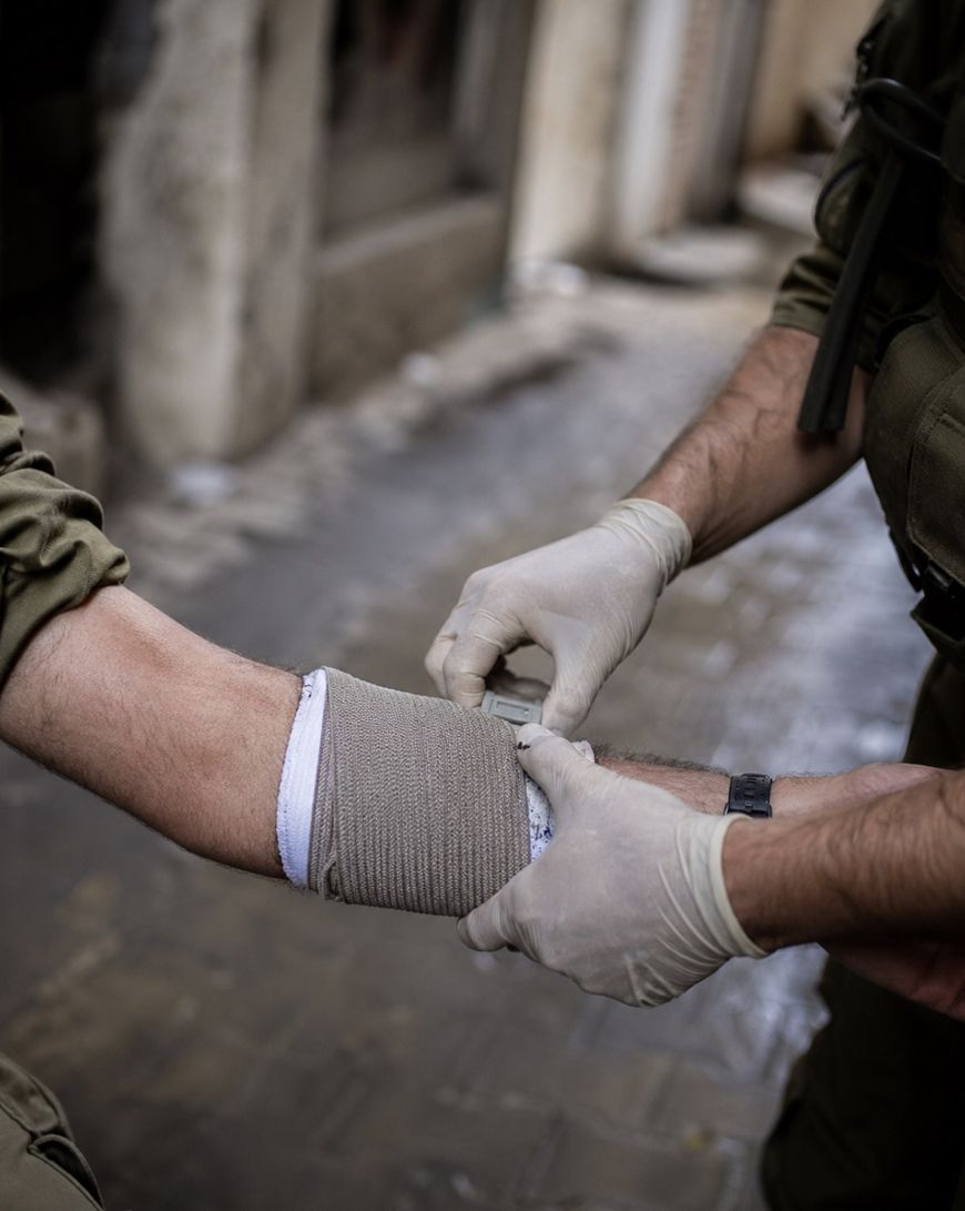 Two people wearing uniforms and latex gloves are applying a bandage to a persons forearm on a street with stone pavement. The scene suggests first aid or medical assistance being given outdoors.