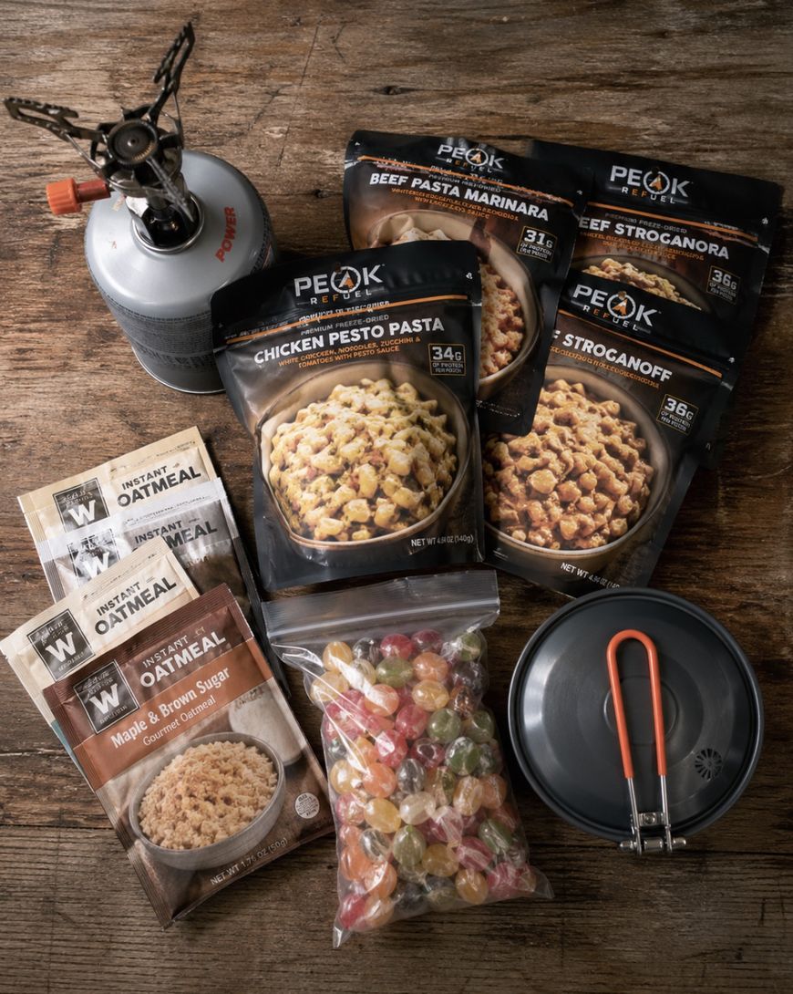 A camping meal setup with a portable stove, a pot, packets of instant oatmeal, three Peak Refuel freeze-dried meal pouches, and a bag of assorted hard candies arranged on a wooden surface.