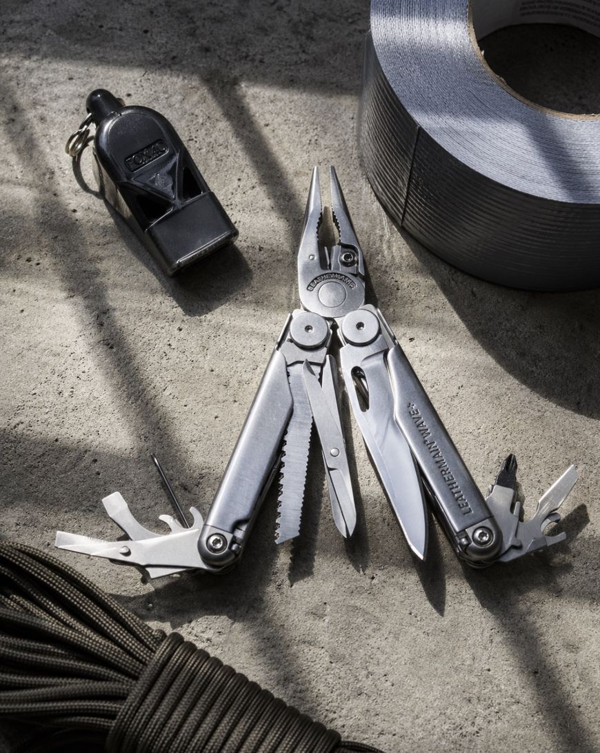 A Leatherman multitool with multiple implements open lies on a concrete surface, surrounded by a black whistle, a roll of gray duct tape, and a coiled black paracord.