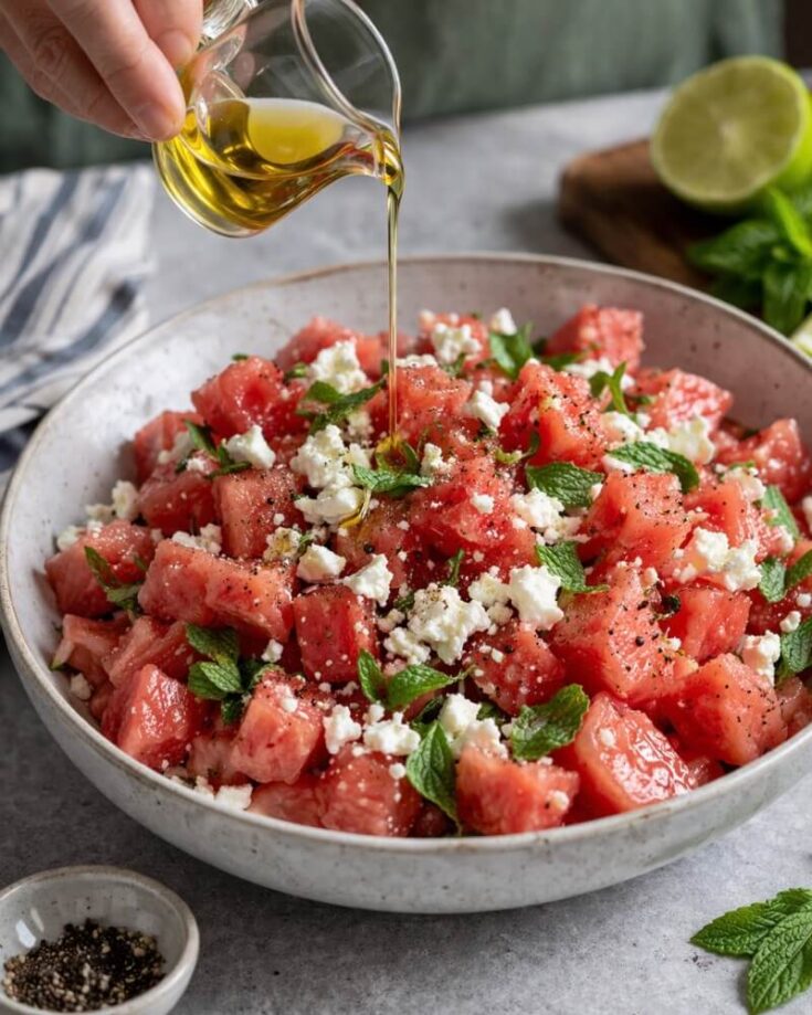 A bowl of watermelon cubes topped with crumbled feta cheese, fresh mint leaves, and black pepper, with olive oil being poured over the salad. A sliced lime, mint, and a small bowl of pepper are nearby.