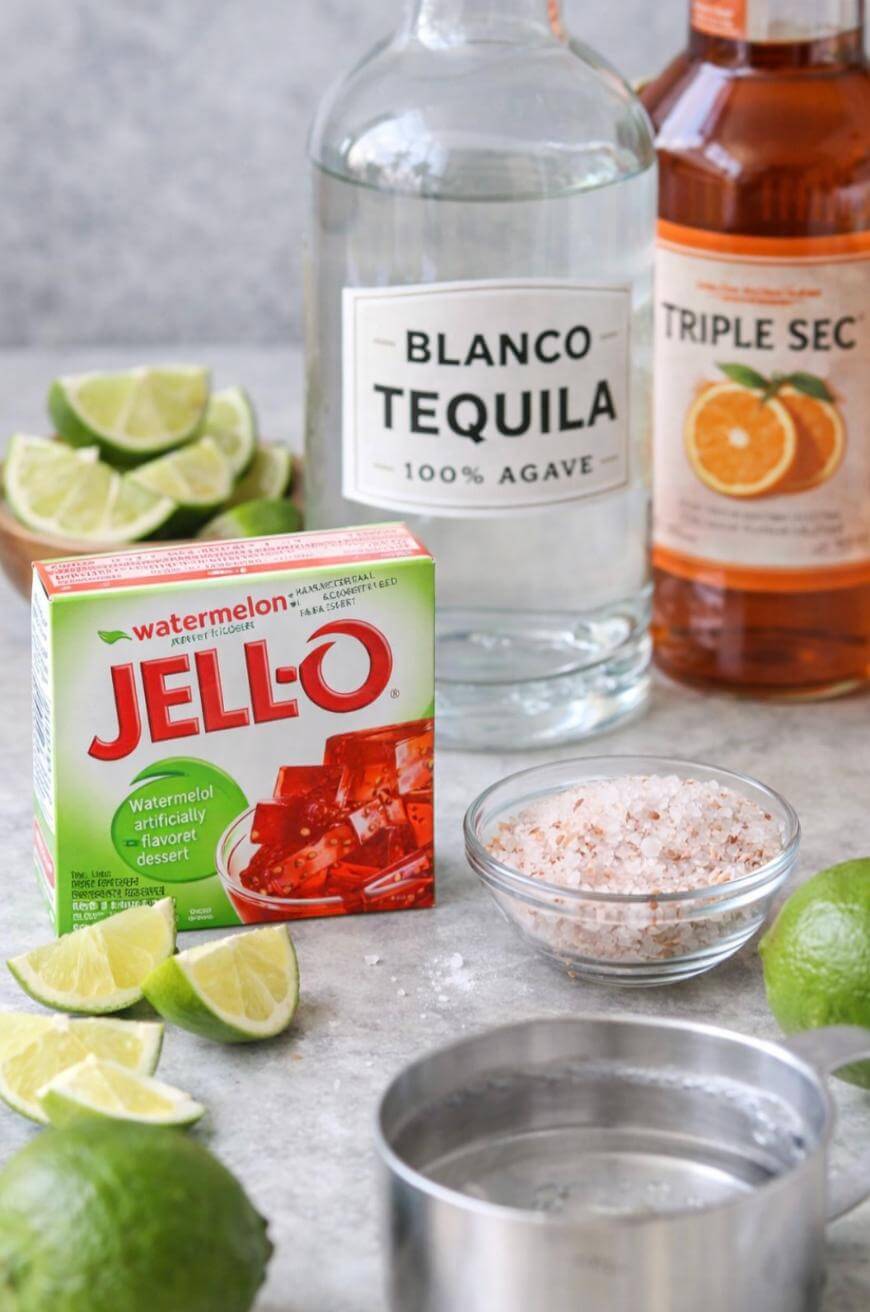 A box of watermelon Jell-O, a bottle of blanco tequila, a bottle of triple sec, a bowl of salt, a metal cup, and sliced limes arranged on a counter.