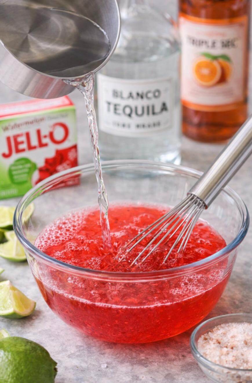 A clear liquid is being poured into a bowl of red gelatin mixture with a whisk inside. Surrounding the bowl are a lime, Jell-O packet, salt, tequila bottle, triple sec bottle, and lime wedges.