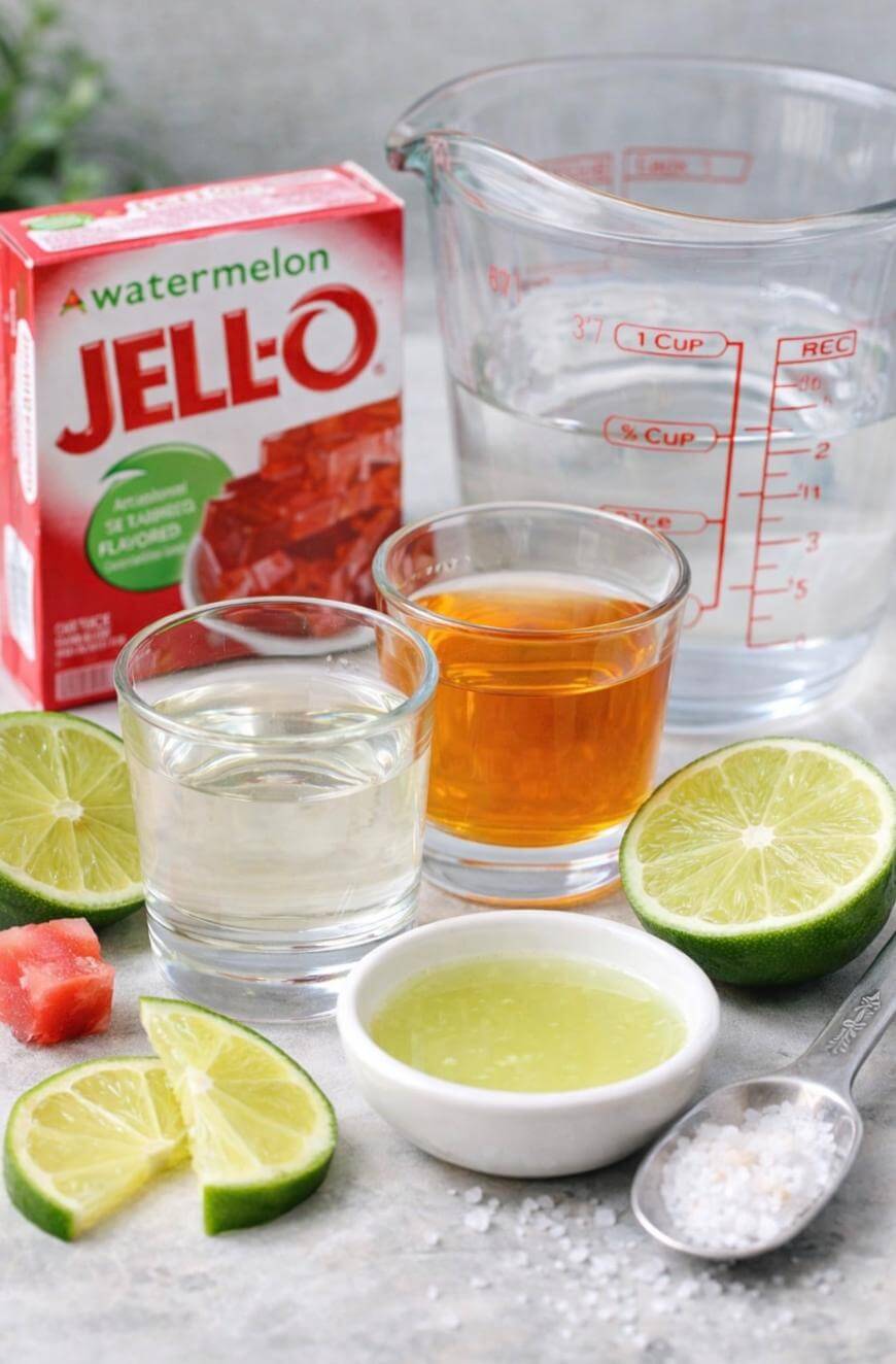 A box of watermelon Jell-O, a measuring cup with water, two glasses with clear and amber liquids, lime slices, a small bowl of lime juice, a spoon with salt, and a watermelon cube arranged on a countertop.