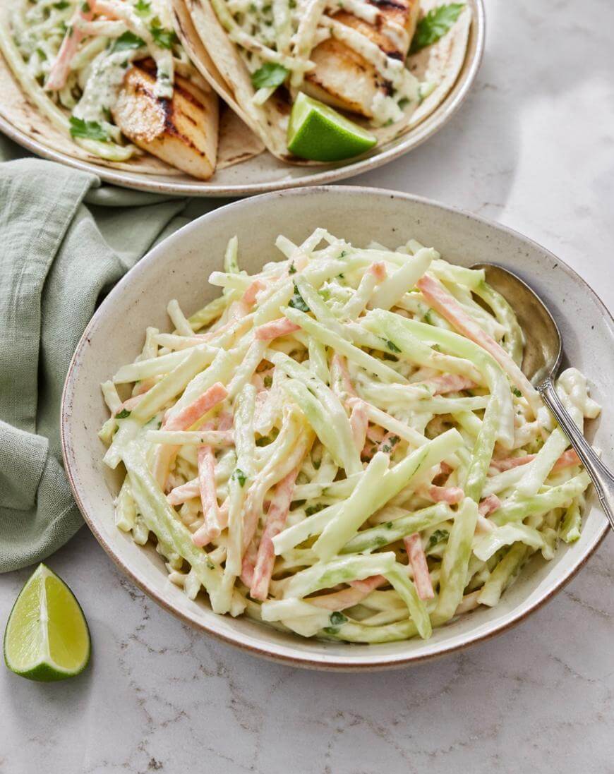 A bowl of creamy coleslaw with shredded cabbage and carrots sits on a marble surface beside grilled chicken tacos topped with slaw and a wedge of lime, with a green cloth napkin nearby.