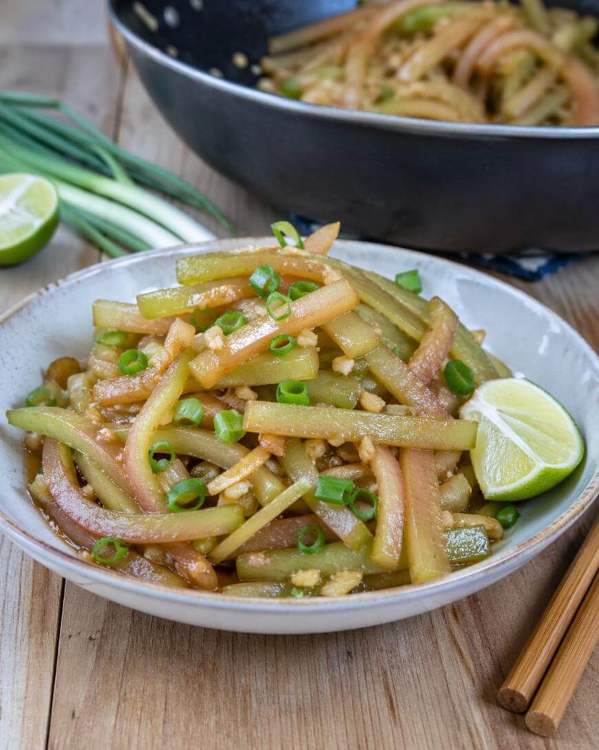 A bowl of stir-fried vegetables garnished with chopped green onions and a lime wedge, with chopsticks and whole green onions beside the bowl on a wooden table. A pan of more vegetables is in the background.
