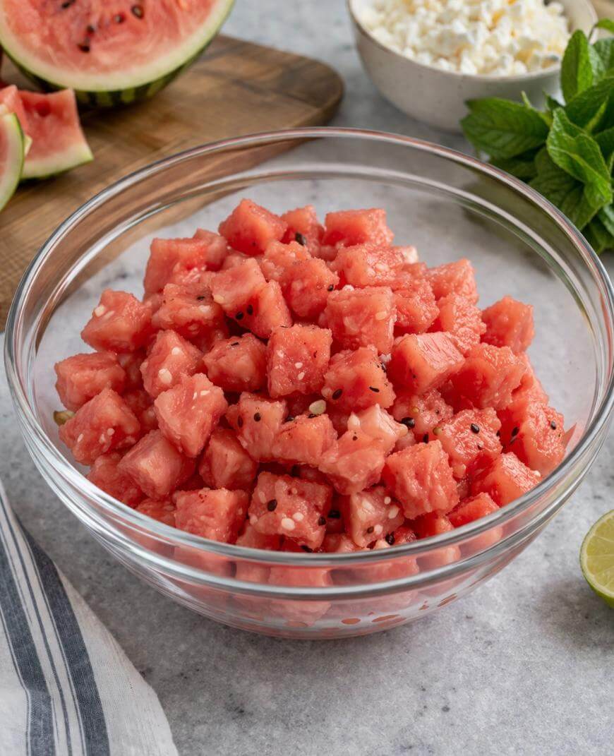 A glass bowl filled with cubed watermelon sits on a countertop. In the background, there are watermelon slices, a bowl of feta cheese, fresh mint leaves, and part of a lime.