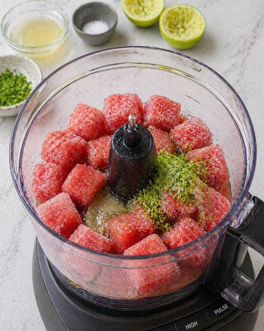 Cubed watermelon, lime zest, and sugar are in a food processor bowl. In the background, there are squeezed lime halves, a bowl of lime juice, and a bowl of zest on a light countertop.