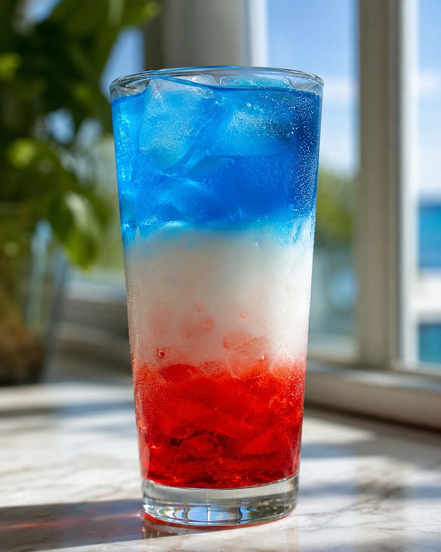 A tall glass filled with a layered drink of blue, white, and red colors over ice, sitting on a marble surface with sunlight coming through a window in the background.