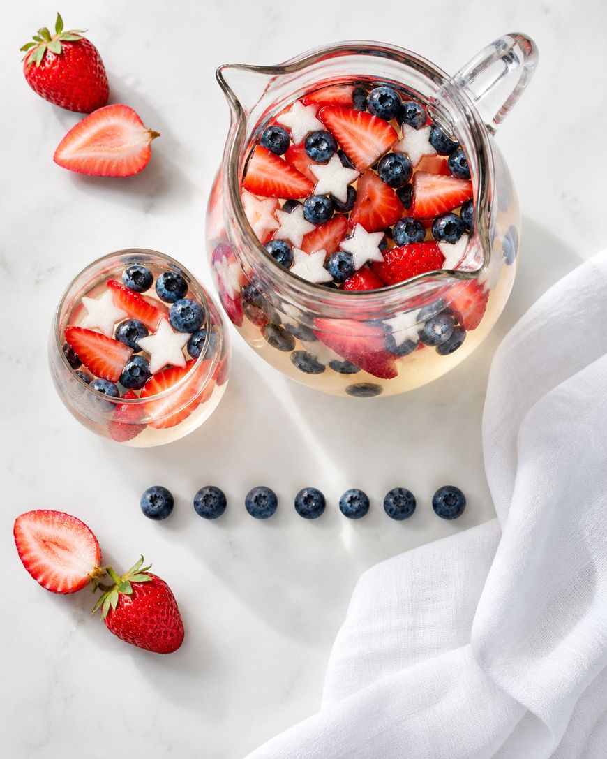 A glass pitcher and glass of fruit-infused water with sliced strawberries, blueberries, and star-shaped pieces of fruit on a white surface, next to fresh berries and a white cloth.