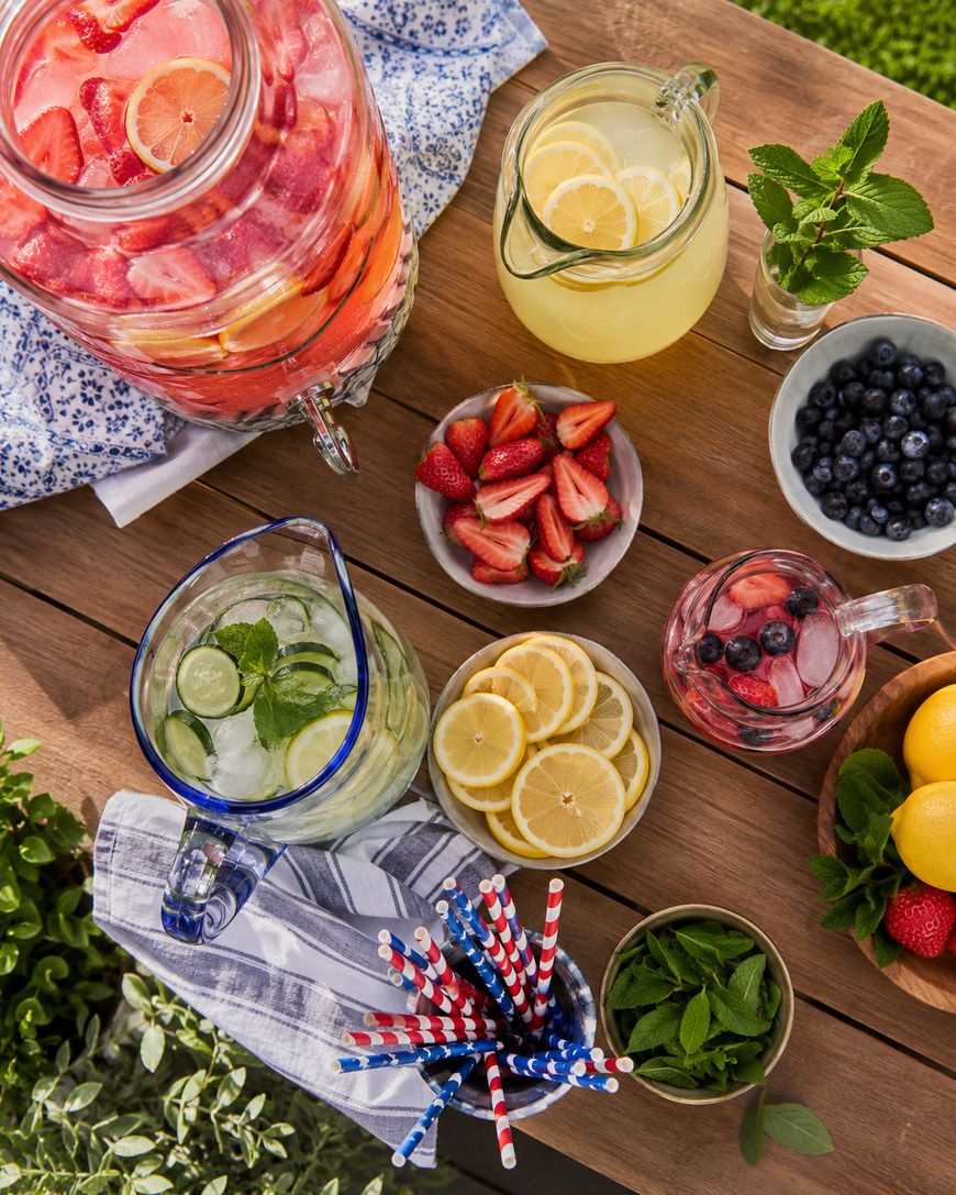 A wooden table set outdoors with pitchers of infused water and lemonade, bowls of fresh strawberries, blueberries, lemon slices, mint, and a container of striped straws. Sunlight highlights the colorful display.