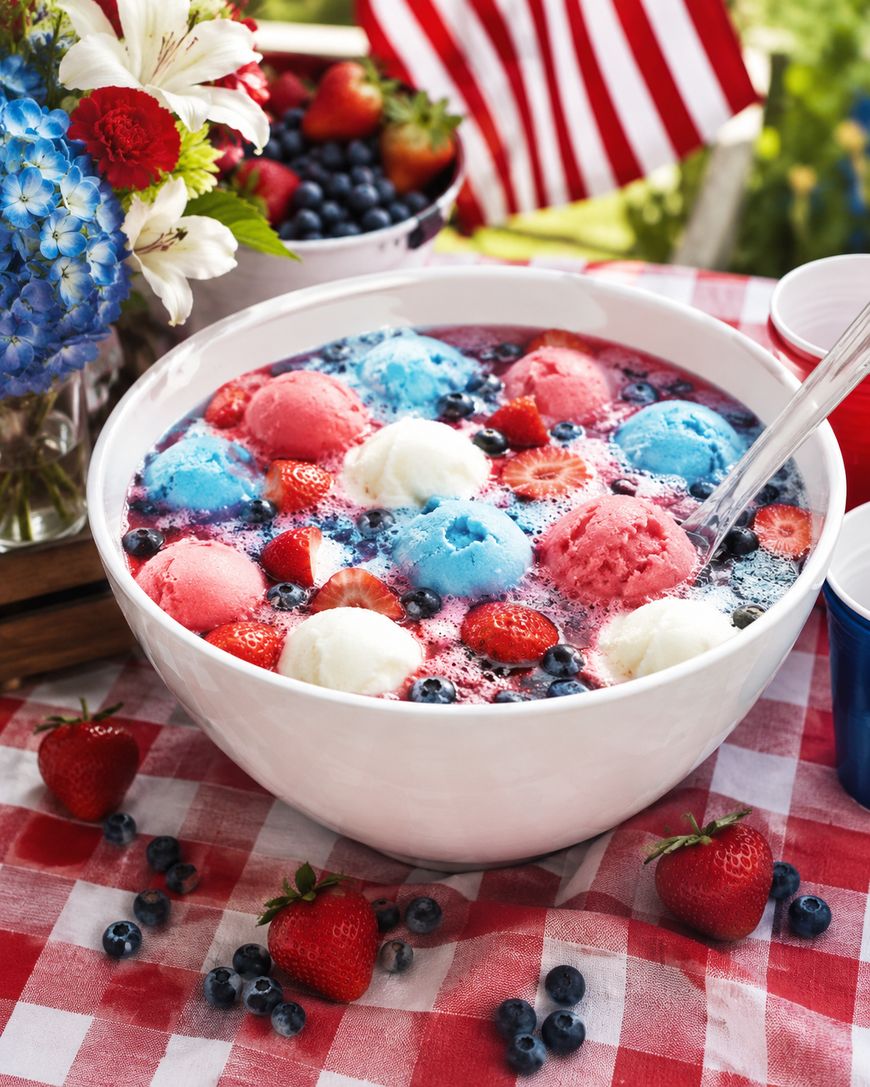 A white bowl filled with red, white, and blue sherbet floats in a fizzy drink, garnished with berries. The bowl sits on a red gingham tablecloth with fresh fruit and flowers nearby, and an American flag in the background.
