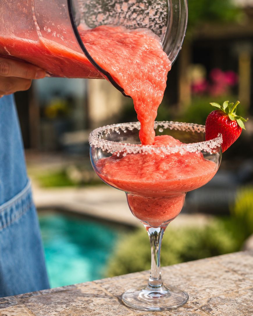 A person pours a thick, pink frozen strawberry drink into a sugar-rimmed margarita glass garnished with a fresh strawberry, outdoors by a pool.