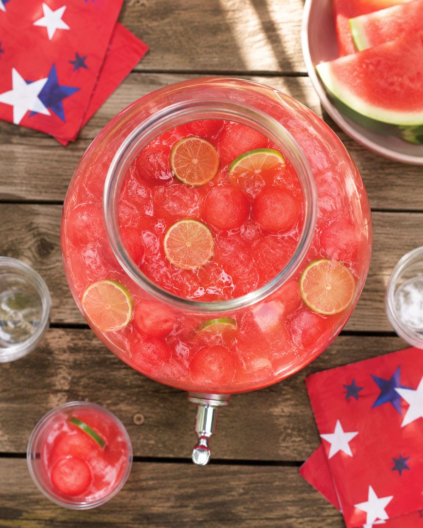 A large glass drink dispenser filled with a pink beverage, watermelon balls, ice, and lime slices sits on a wooden table, surrounded by glasses, red star-patterned napkins, and watermelon slices.