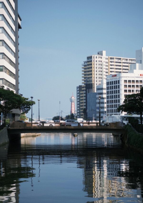 A calm urban canal passes under a bridge, flanked by modern buildings. In the distance, a red-and-white tower rises against a clear blue sky, reflecting in the water below.