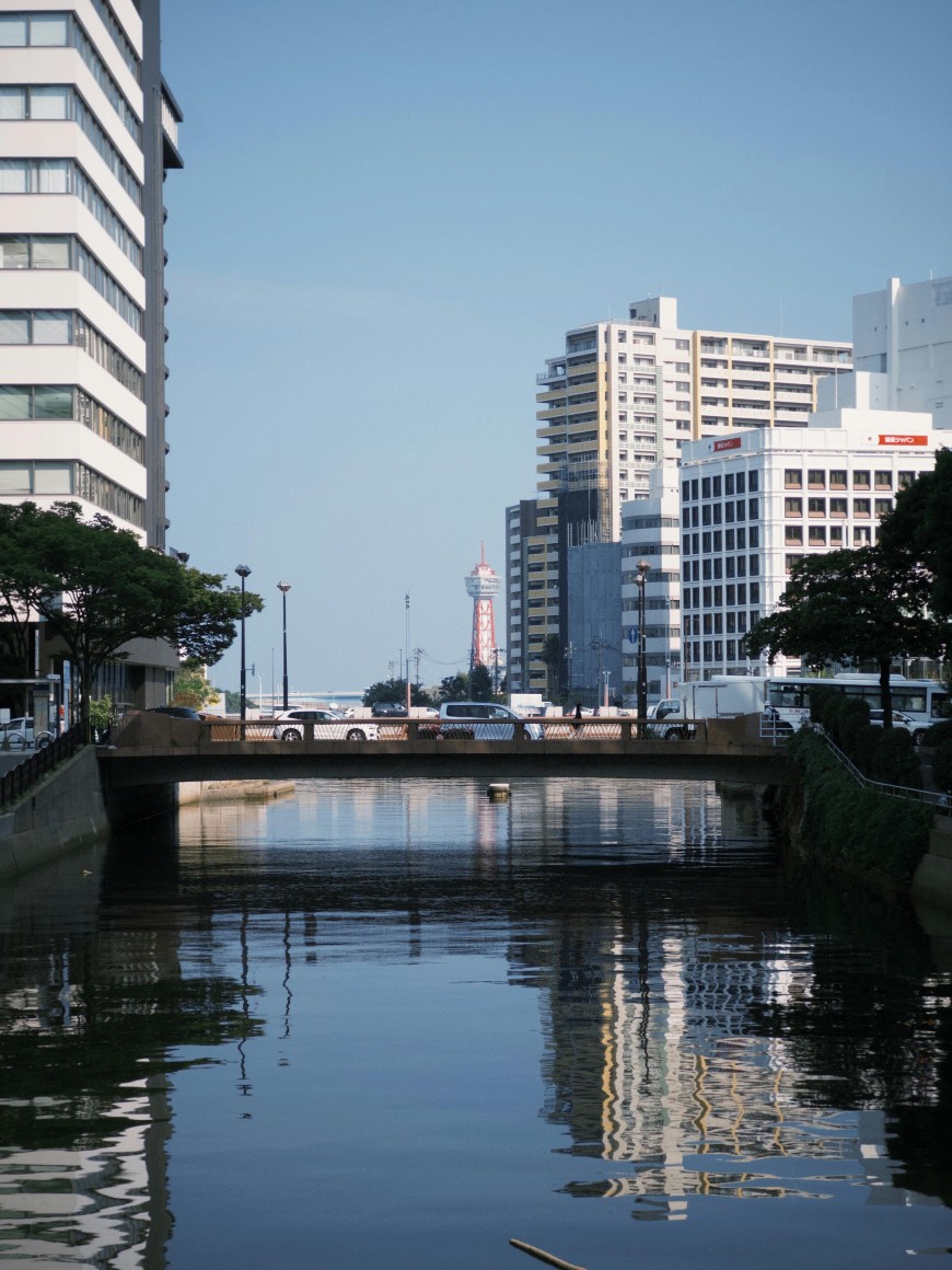 A view of a city canal with a small bridge crossing over it, surrounded by tall modern buildings. Trees line the water, and a red and white observation tower is visible in the background under a clear blue sky.