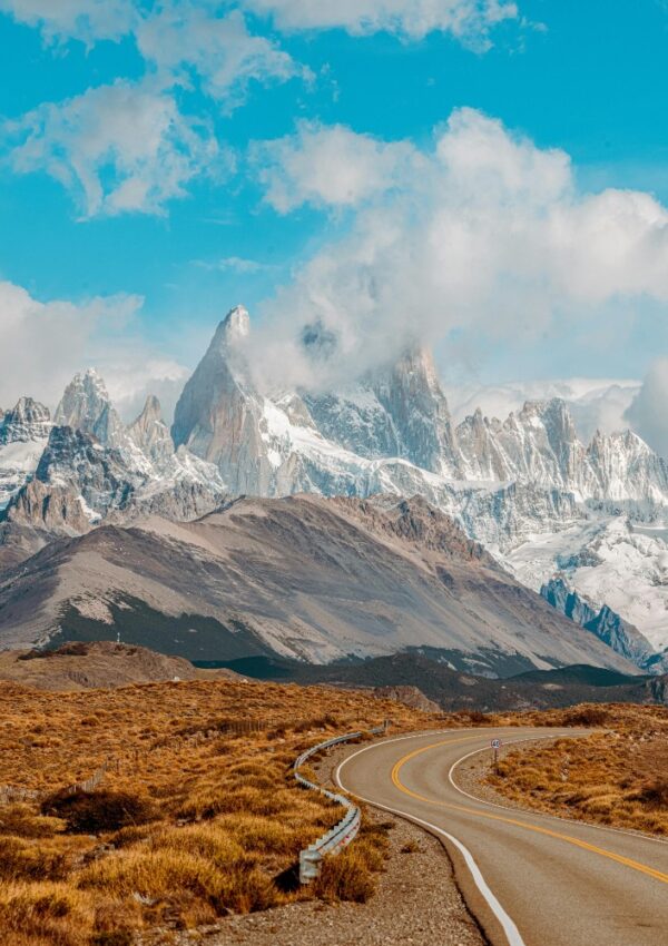 A winding road leads toward snow-capped jagged mountains under a bright blue sky with scattered clouds, surrounded by brown and yellow grassy plains.