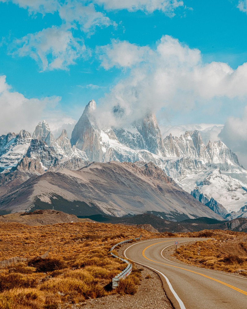 A winding road leads toward snow-capped jagged mountains under a bright blue sky with scattered clouds, surrounded by brown and yellow grassy plains.