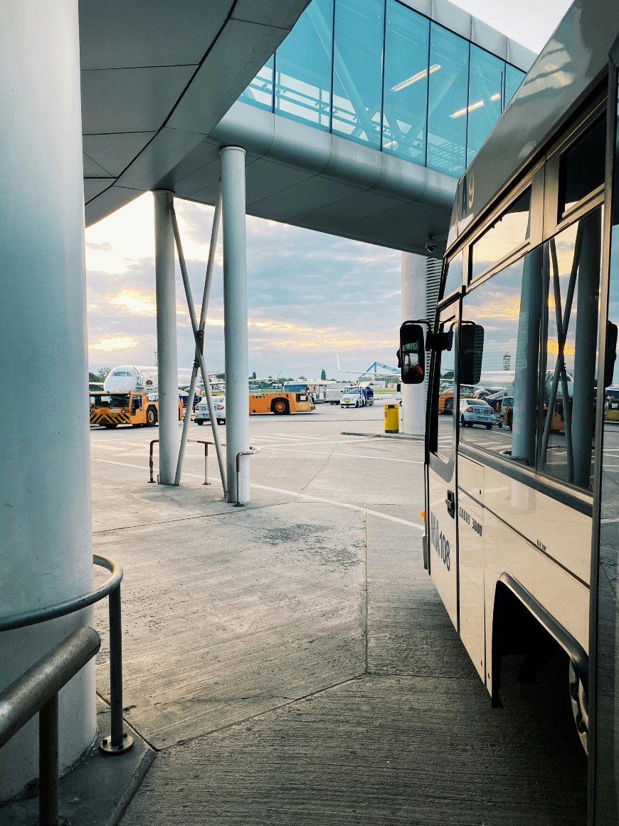 A bus is parked at an airport terminal with a covered walkway above. Airplanes, airport vehicles, and cars are visible in the background under a cloudy sky at sunset.