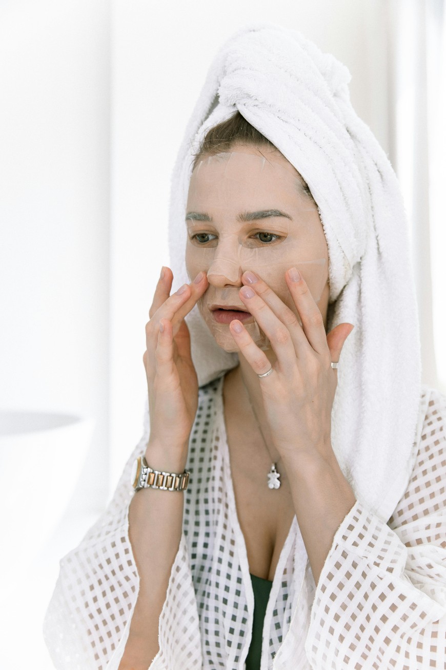 A woman wearing a white bathrobe and towel on her head applies a sheet face mask, gently pressing it onto her face. She stands in a bright bathroom, focusing on her skincare routine.