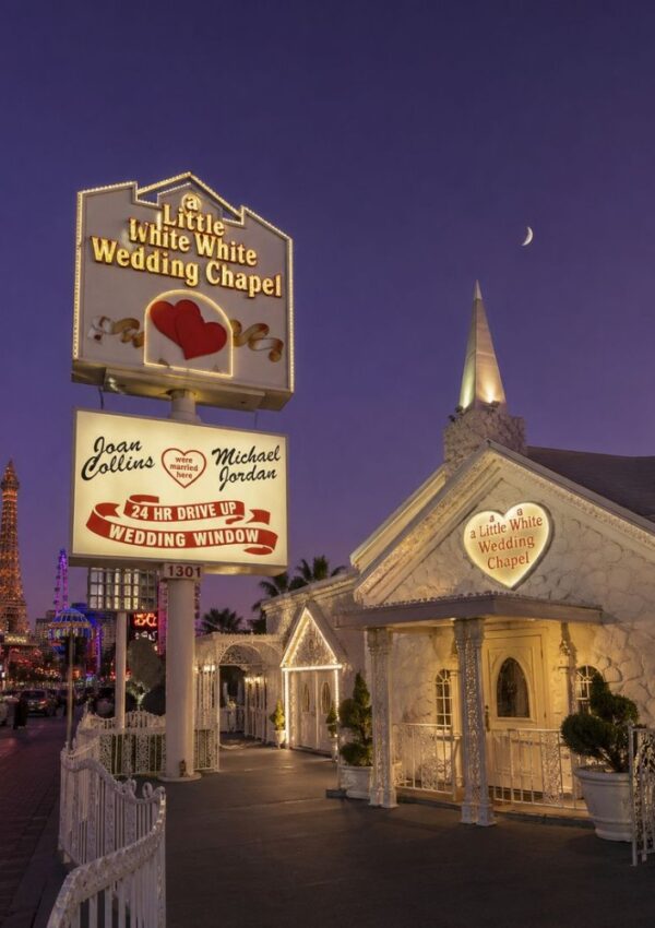 A brightly lit Little White Wedding Chapel in Las Vegas at dusk, with glowing signs, a heart motif, and a crescent moon in the purple sky—an iconic spot perfect for Vegas elopement tips. The Eiffel Tower replica shimmers in the distance.