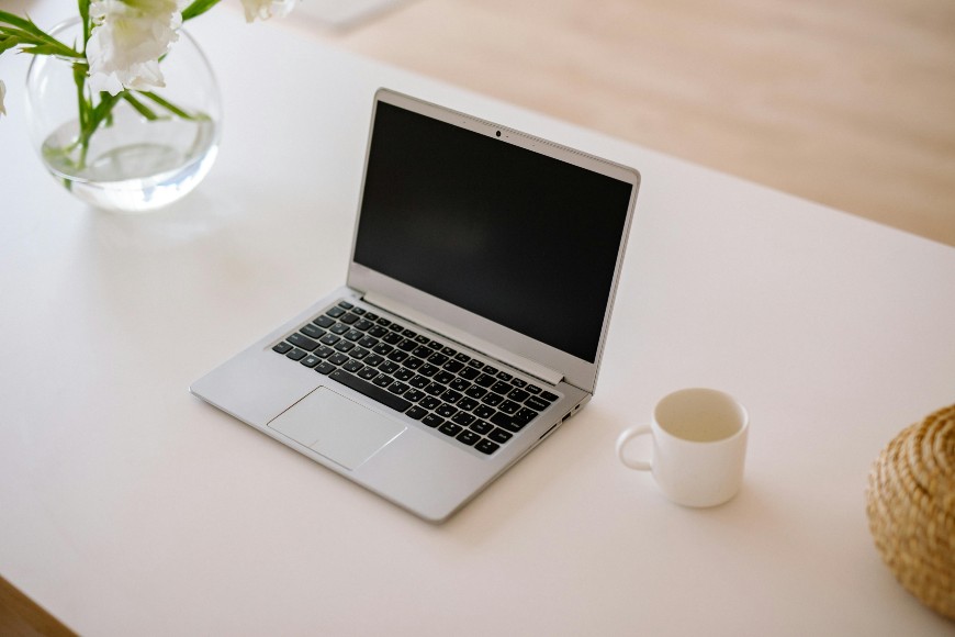 A silver laptop with a black screen sits on a white table beside a white coffee mug, a woven item, and a glass vase with white flowers. The scene is well-lit with minimal, modern decor.