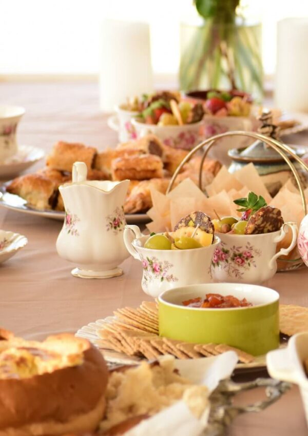 A table set for tea with floral china, various pastries, crackers, fruit, a cream pitcher, and small bowls of jams and spreads, all arranged on a pastel tablecloth.