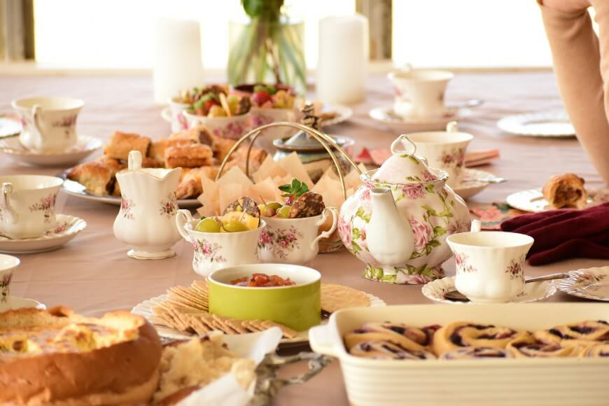 A table set for tea with floral china, a teapot, cups, plates of pastries, crackers, fruit, and a casserole dish. The scene is bright and inviting, with a soft tablecloth and a relaxed, elegant atmosphere.