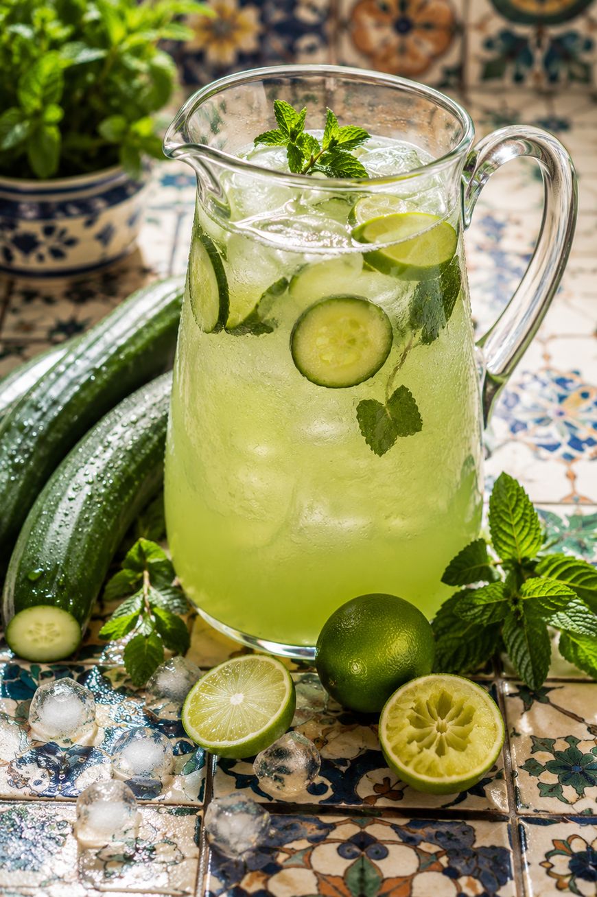 A glass pitcher filled with cucumber limeade, garnished with cucumber slices, lime slices, and fresh mint. Whole cucumbers, limes, mint leaves, and ice cubes are arranged on a patterned tile surface.