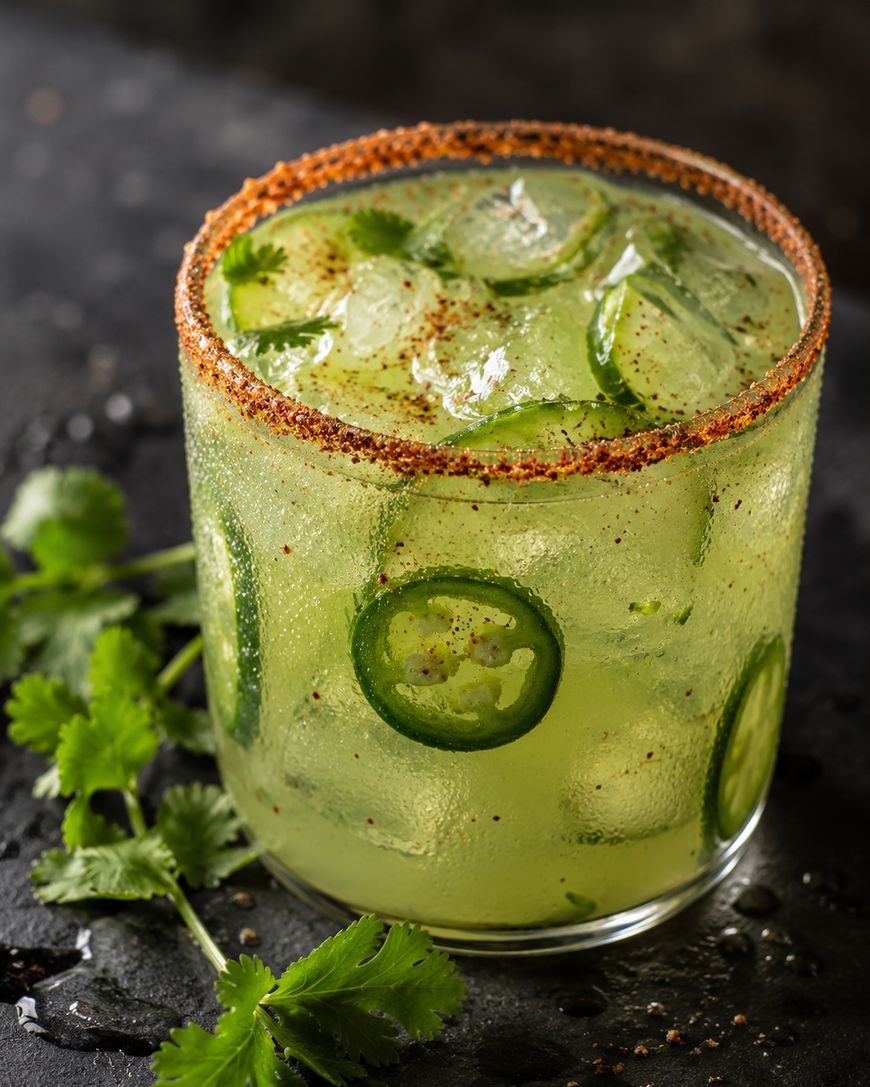 A glass of green cocktail with ice cubes, sliced jalapeños, and a chili powder rim, garnished with fresh cilantro on a dark surface.