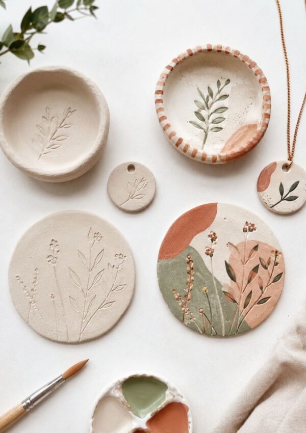 Handcrafted ceramic dishes and pendants with delicate botanical designs in earthy tones, arranged on a white surface—perfect inspiration for air dry clay ideas—with a paintbrush, pastel palette, and green leaves nearby.