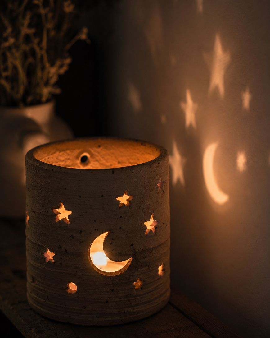 A ceramic candle holder with star and crescent moon cutouts casts glowing star and moon shapes onto a nearby wall in a dimly lit room. A blurred vase with dried flowers is visible in the background.