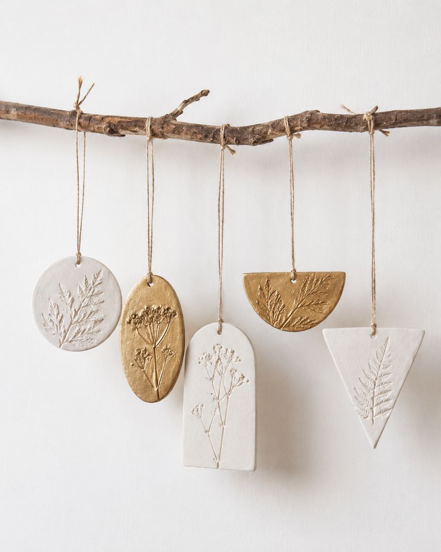 Five botanical clay ornaments, each with pressed plant designs, hang from a rustic branch by twine. The ornaments are in oval, round, rectangle, half-circle, and triangle shapes in white and gold tones.
