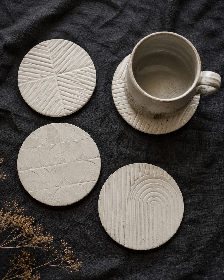 Four round, textured ceramic coasters with different geometric patterns are arranged on a dark fabric surface; one coaster has a ceramic mug on it. Dried flowers are visible in the bottom left corner.