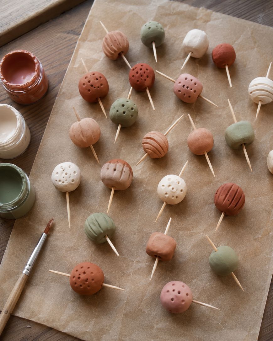 Round clay beads in earth tones are skewered on toothpicks and arranged on parchment paper. Paint jars and a brush are nearby, with some beads painted and textured with holes or lines.