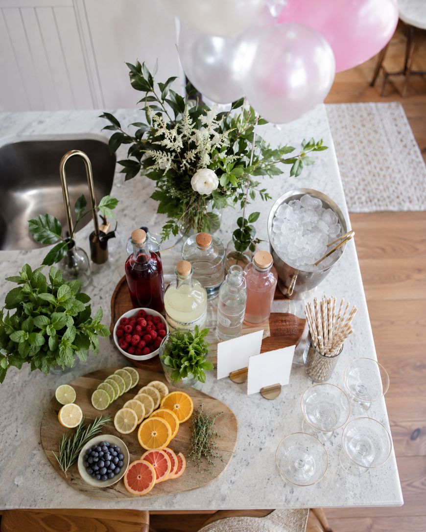 A marble countertop with sliced citrus fruits, berries, herbs, syrups, glasses, an ice bucket, and bottles. Balloons and a floral arrangement decorate the space, creating a festive, refreshing drink station.