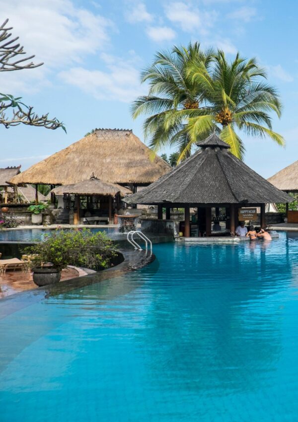 A tropical resort pool with clear blue water, surrounded by thatched-roof huts and palm trees. Two people are sitting at a swim-up bar, and lush greenery decorates the area under a blue sky.
