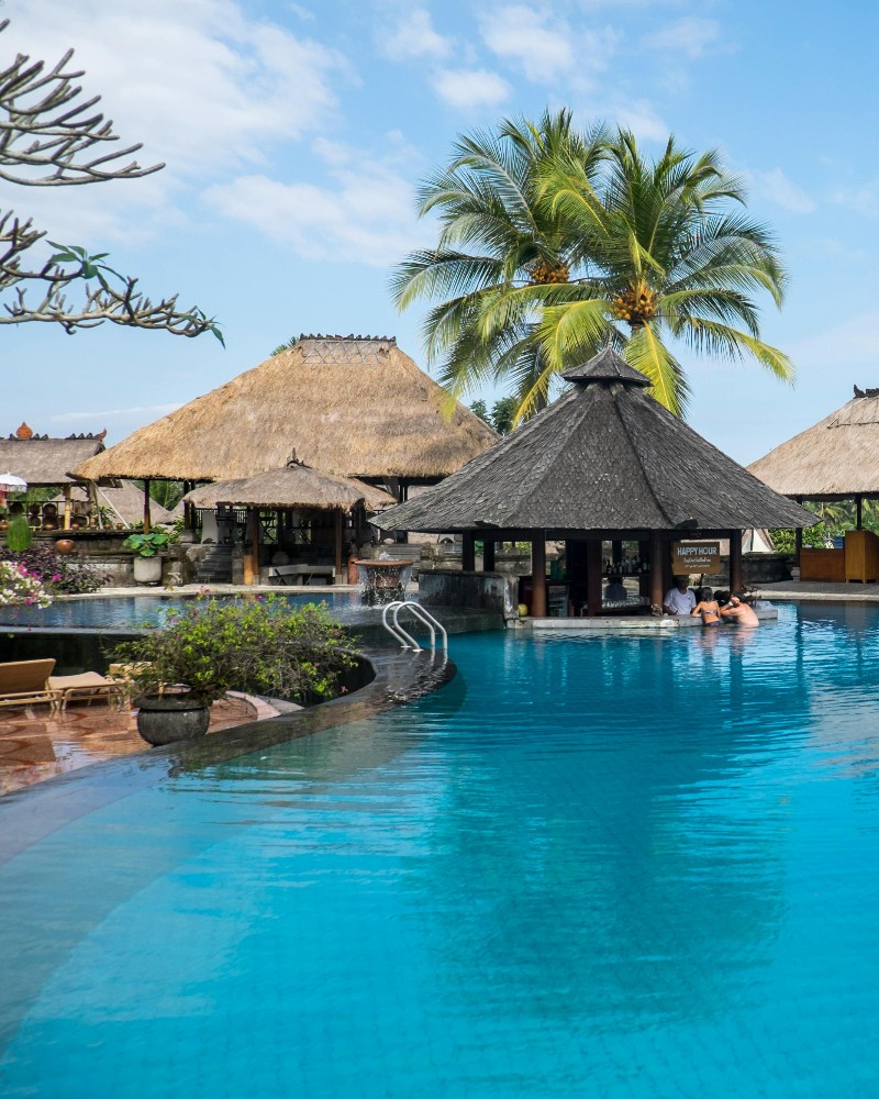 A tropical resort pool with clear blue water, surrounded by thatched-roof huts and palm trees. Two people are sitting at a swim-up bar, and lush greenery decorates the area under a blue sky.