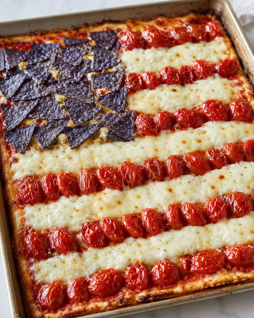 A rectangular sheet pan pizza decorated like the American flag, with rows of melted cheese and cherry tomatoes for stripes, and blue corn tortilla chips in the top left corner to represent stars.