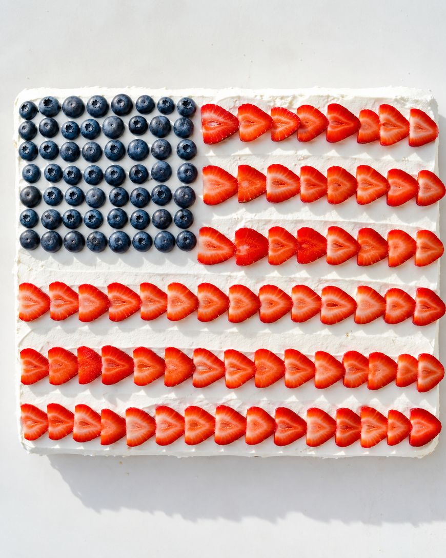 A rectangular cake decorated to look like an American flag with blueberries forming the top left corner and rows of sliced strawberries over white frosting creating red and white stripes.