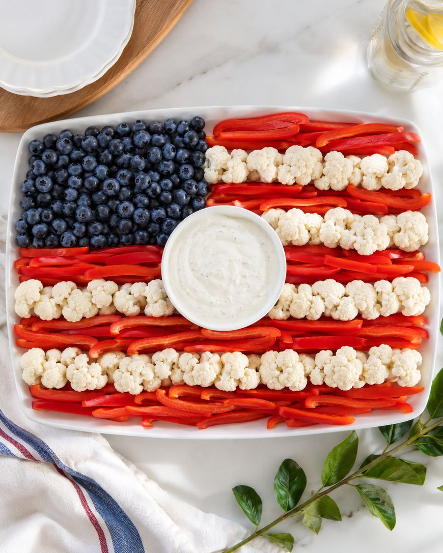 A rectangular platter arranged like the American flag with blueberries as the blue section, red bell pepper strips as red stripes, cauliflower as white stripes, and a bowl of ranch dip in the center.