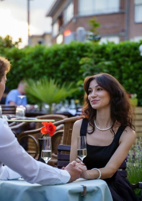 A couple sits at an outdoor restaurant table, holding hands and smiling at each other. The woman wears a black dress and pearl necklace; a red flower and glasses of champagne are on the table. Greenery and buildings appear in the background.