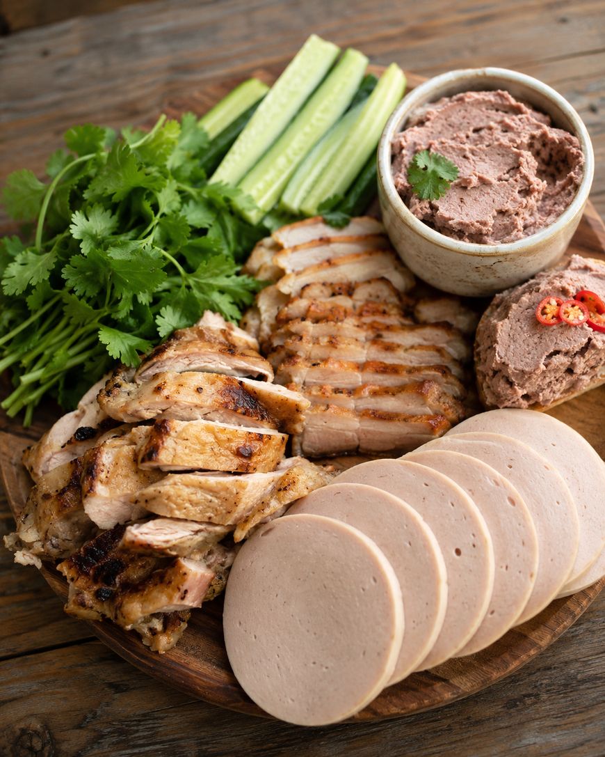 A wooden platter with sliced grilled chicken, slices of pork sausage, cucumber sticks, fresh cilantro, and a bowl of pâté garnished with cilantro and red chili, arranged on a rustic wooden table.
