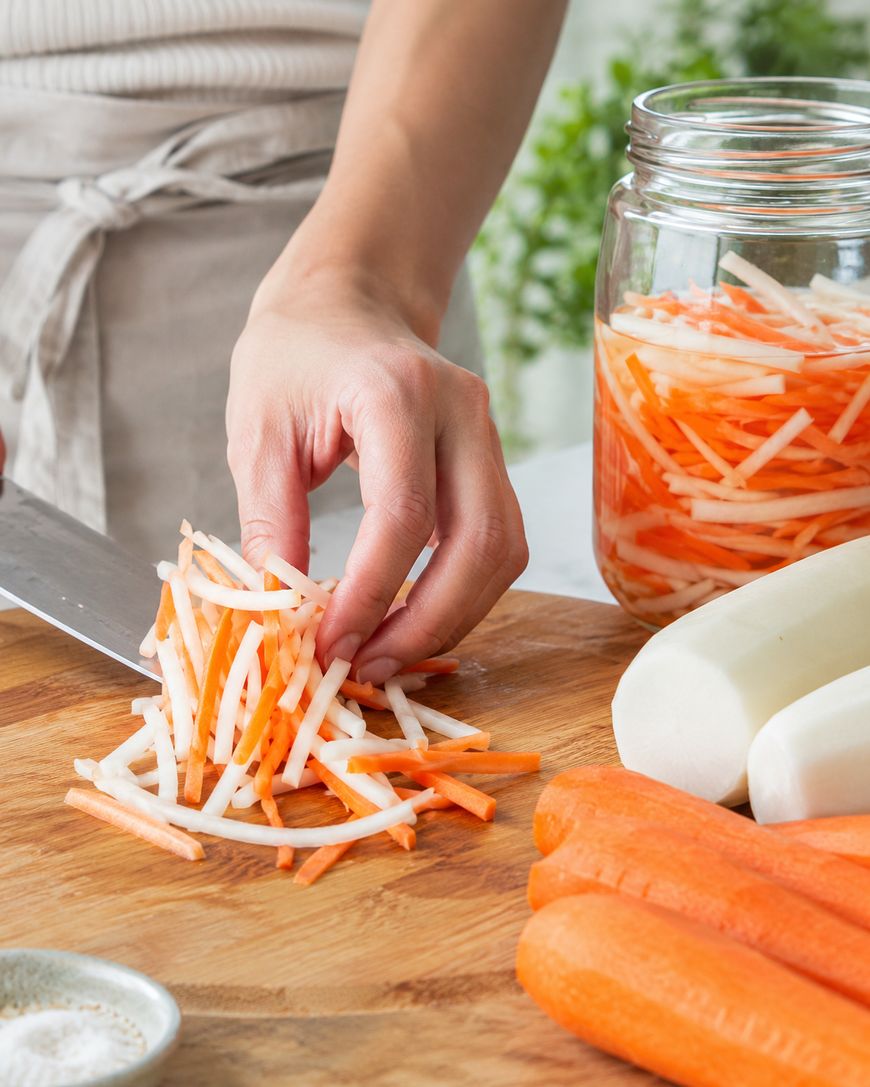 A person’s hand arranges thinly sliced white and orange vegetable strips, likely daikon and carrot, on a cutting board. A jar filled with similar strips is nearby, along with whole carrots and daikon.