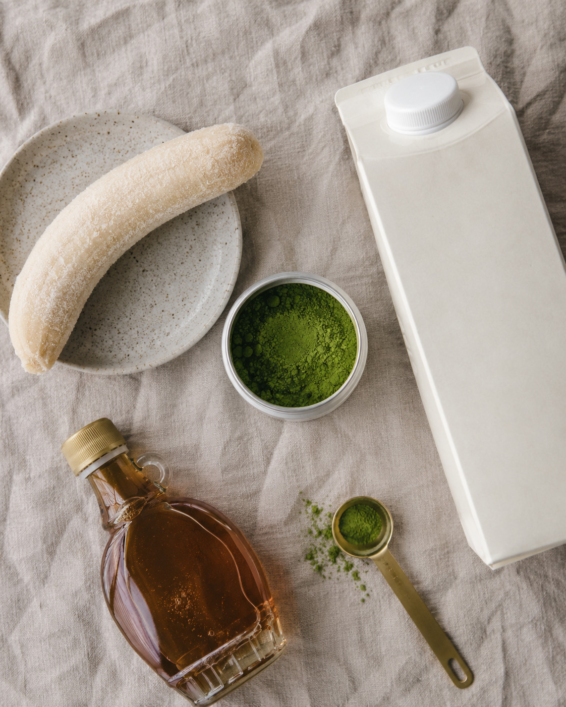 A frozen banana on a plate, a carton of milk, a small jar and measuring spoon of green matcha powder, and a glass bottle of maple syrup are arranged on a light linen surface.